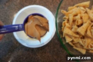 A measuring cup filled with peanut butter is held above a white container, ready to create some unicorn horn muddy buddies. Next to it, a green bowl holds a pile of rolled corn snacks, resting on the brown countertop in perfect harmony.