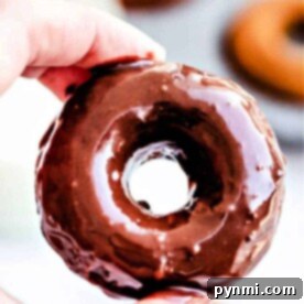Close-up of a hand holding a glazed donut with blurred baked pumpkin donuts in the background.