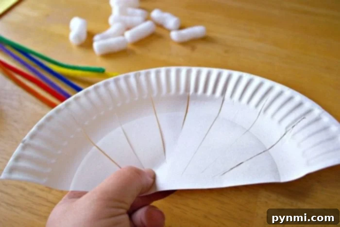 Kaleidoscope Weave Art 3 A person's hand is holding a cut white paper plate, showcasing multiple slits for rainbow weaving. Colorful pipe cleaners and white packing peanuts are scattered on a wooden surface in the background, adding to the playful art scene.