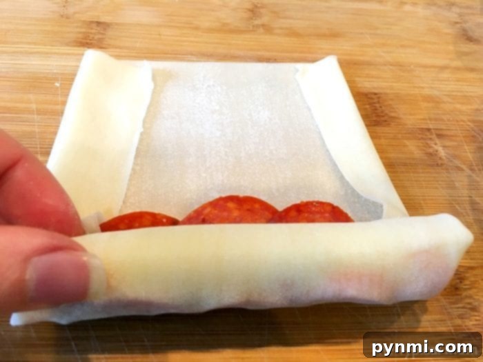 Pizzarolls 5 A hand demonstrates rolling a pizza egg roll with pepperoni and cheese on a wooden surface, showing the preparation step.