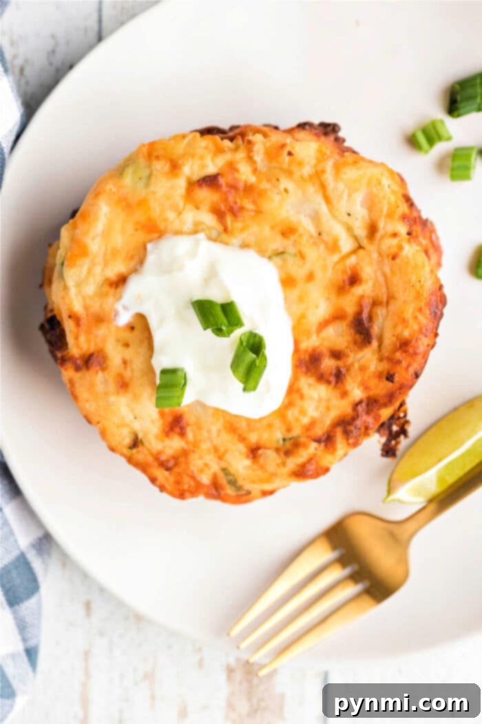 A cheesy potato pancake topped with sour cream and chopped green onions, served with lime wedges on a white plate, reminiscent of fried mashed potatoes with cheese.