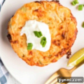 A cheesy potato pancake topped with sour cream and chopped green onions, served with lime wedges on a white plate, reminiscent of fried mashed potatoes with cheese.