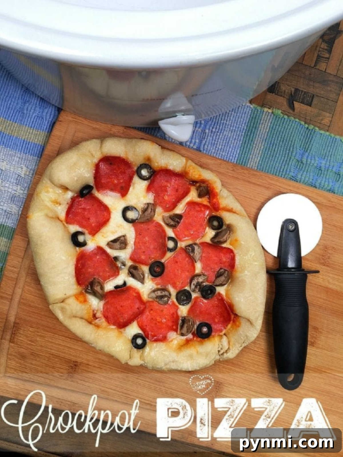 A homemade crock pot pizza with pepperoni, mushrooms, and olives rests on a cutting board next to a pizza cutter and crockpot.