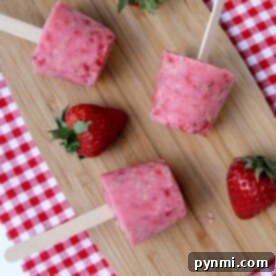 Four Fresh Fruit Frozen Yogurt Popsicles rest on a wooden board with fresh strawberries, set atop a red gingham cloth.