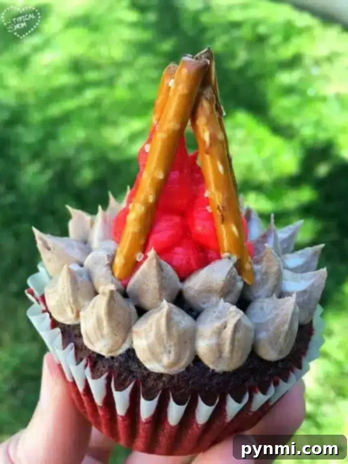 A captivating close-up of a beautifully decorated camping cupcake, expertly designed to look like a miniature campfire. It features intensely vibrant red frosting flames that seem to flicker, sturdy pretzel stick logs perfectly arranged, and a rich chocolate base representing the earth. The cupcake is gently held in a hand against a subtly blurred grassy backdrop, powerfully enhancing the outdoor and natural theme, making it irresistible.