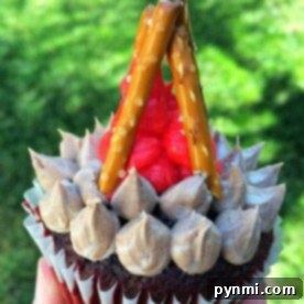 A close-up, top-down view of a beautifully decorated campfire cupcake, featuring realistic pretzel sticks arranged as logs and vibrant red frosting mimicking flickering flames. The rich chocolate base completes the rustic, outdoor theme.