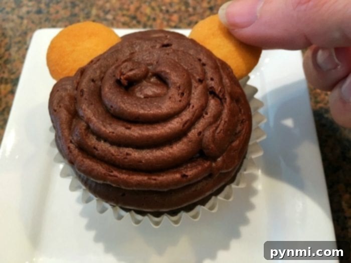 A hand gently places a mini vanilla wafer cookie onto a chocolate cupcake swirled with frosting, forming one of the monkey's charming ears, showcasing the ease of creation.