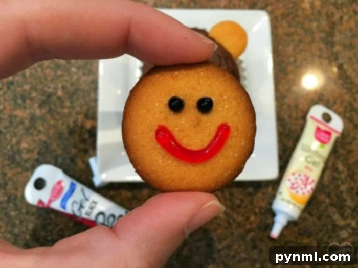 A hand holding a vanilla wafer cookie decorated with a cheerful red smile and two black gel dots for the nose, with colorful icing tubes in the background, demonstrating a key step for monkey or bear cupcakes.