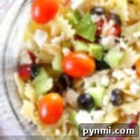 A vibrant Mediterranean pasta salad features a close-up of bowtie pasta adorned with cherry tomatoes, crisp cucumbers, briny olives, feta cheese, and fresh green peppers.