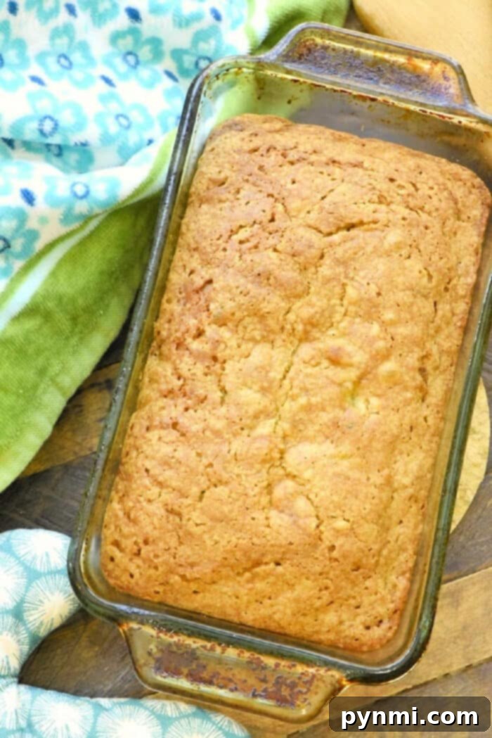 A perfectly baked loaf of zucchini pineapple bread in a glass pan, resting on a wooden surface with patterned towels nearby, highlighting its golden-brown crust.