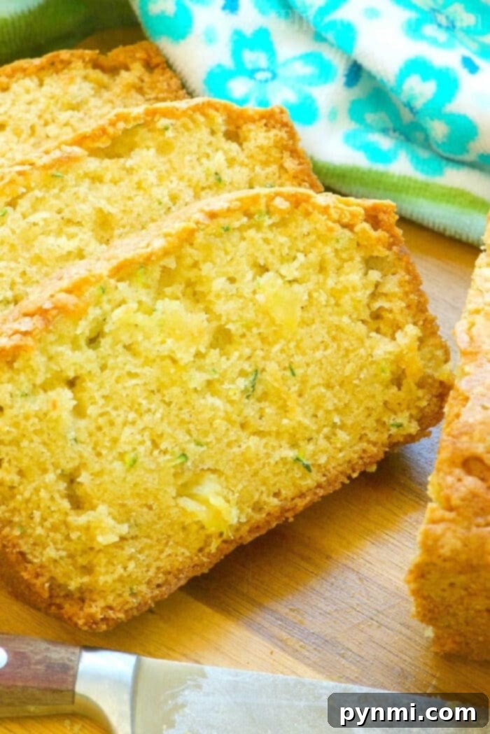 Beautifully displayed slices of zucchini pineapple bread on a wooden surface, showing a golden crust and a moist interior with visible shreds of zucchini and pineapple bits. A patterned green and white cloth and a knife are in the background.