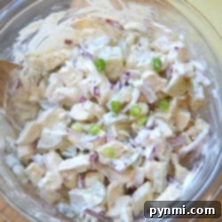 A glass bowl brimming with creamy rotisserie chicken salad, featuring diced onions, peas, and a medley of flavors sits enticingly on a wooden board.