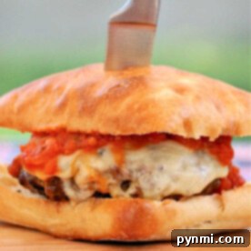 A New York pizza burger with melted cheese and tomato sauce on a bun, topped with a knife, sits enticingly on a wooden board.