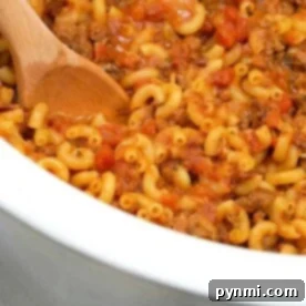 A wooden spoon stirs a pot of macaroni with ground meat and tomato sauce, reminiscent of a homemade crockpot hamburger helper.