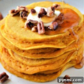 A stack of pumpkin pancakes, one of the easy pumpkin desserts, topped with butter, pecans, and drizzled maple syrup sits on a plate. A few pecans are scattered around the plate. The pancakes have a light golden-brown color.