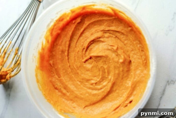 A bowl of creamy pumpkin cake mix with a whisk sits on a white marble countertop, ready to transform into delightful mini bundt cakes.