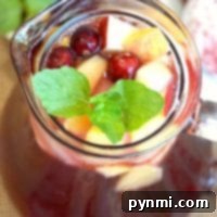 A glass pitcher of sparkling cranberry sangria brimming with apple cubes, fresh cranberries, and a mint garnish.