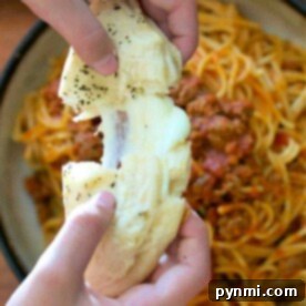 Hands pulling apart cheesy garlic bread over a plate of spaghetti with meat sauce, using flaky layers reminiscent of a refrigerated biscuit filled with cheese.
