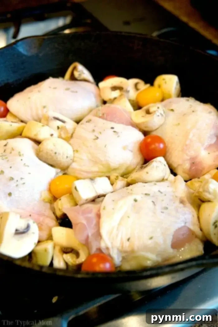 Baked bone in chicken thighs recipe that is made in one pot! So easy and done from start to finish in just 30 minutes with only 5 ingredients. Delicious. A cast iron skillet on a stove contains raw chicken thighs, quartered mushrooms, and cherry tomatoes, ready for cooking. While you prep your ingredients, you might wonder, *How long to bake bone-in chicken thighs at 350*? Seasoned with herbs, this dish promises a flavorful experience.