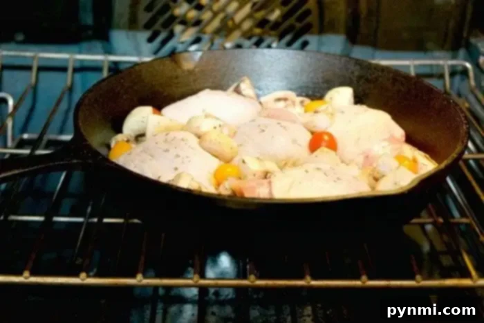 Perfectly Baked Bone-in Chicken Thighs 3 A cast iron skillet containing raw chicken pieces, cherry tomatoes, and mushrooms is in an oven with the door closed. The oven's heating elements are visible, and the food appears to be seasoned with herbs. For those wondering how long to bake bone-in chicken thighs at 350, this setup offers a savory answer.