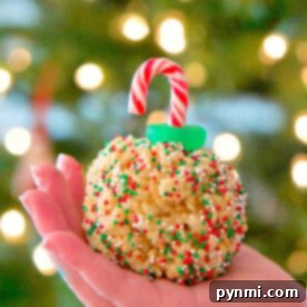 A hand holds a festive Rice Krispy treat ornament adorned with sprinkles and a candy cane hook, set against the soft glow of a blurred tree.