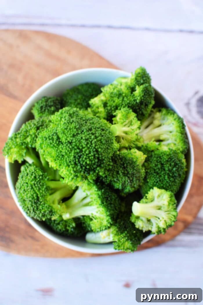A bowl filled with fresh green broccoli florets, perfect for a delicious steamed broccoli recipe, is placed on a wooden cutting board. The background shows a lightly textured white surface.