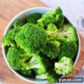 A bowl filled with fresh green broccoli florets, perfect for a delicious steamed broccoli recipe, is placed on a wooden cutting board. The background shows a lightly textured white surface.