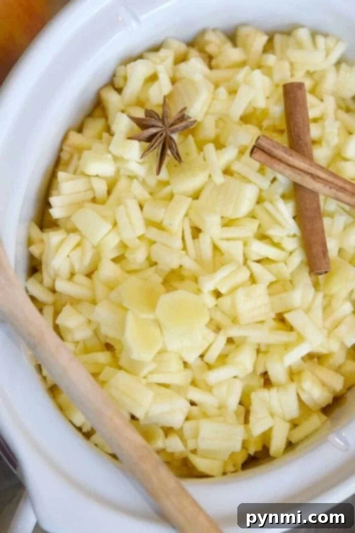 A white bowl filled with diced apples, ready to transform into slow cooker apple butter. A star anise and a cinnamon stick are placed on top of the apples, while a wooden spoon rests on the edge of the bowl.