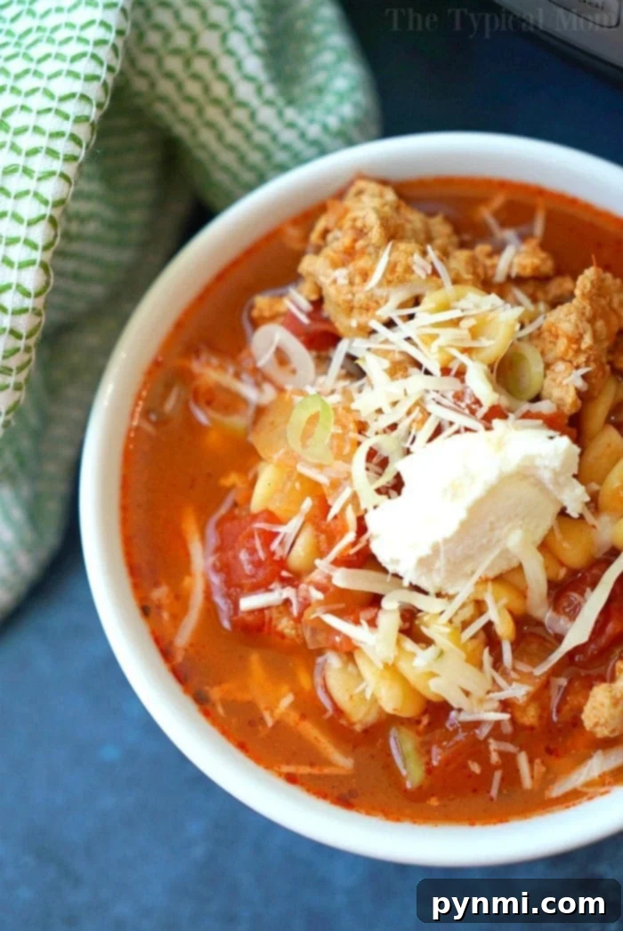 This easy Instant Pot lasagna soup is amazing and healthy!! You can make it in the slow cooker too but in your pressure cooker it's done in just 8 minutes! Your favorite pasta dinner is now a soup and much easier to make. #lasagnasoup #lasagna #soup #instantpot #pressurecooker #easy A close-up of a vibrant bowl of Instant Pot lasagna soup, garnished with grated cheese and a dollop of cream, resembling a hearty minestrone. The soup contains pasta, tomatoes, and broth, with a green and white cloth in the background.