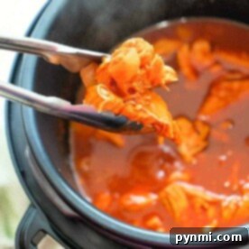 Tongs grasping tender instant pot shredded chicken in a pressure cooker brimming with rich red sauce.