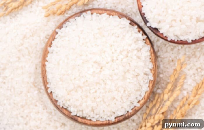 A wooden bowl brimming with long grain white rice rests on a surface sprinkled with rice grains, surrounded by wheat stalks, highlighting the main ingredient.