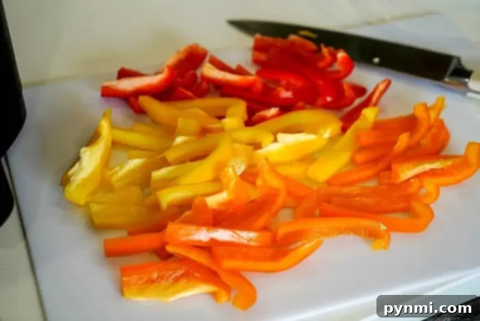 Sliced red, yellow, and orange bell peppers are arranged on a white cutting board, ready for the next step in learning how to blanch bell peppers. A knife is placed nearby on the board.