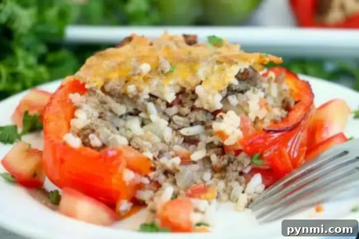 A stuffed red bell pepper, blanched to perfection, sits on a white plate filled with a savory mix of ground meat and rice, topped with melted cheese. Diced tomatoes and chopped herbs are artfully scattered around the pepper, while a fork rests beside it.