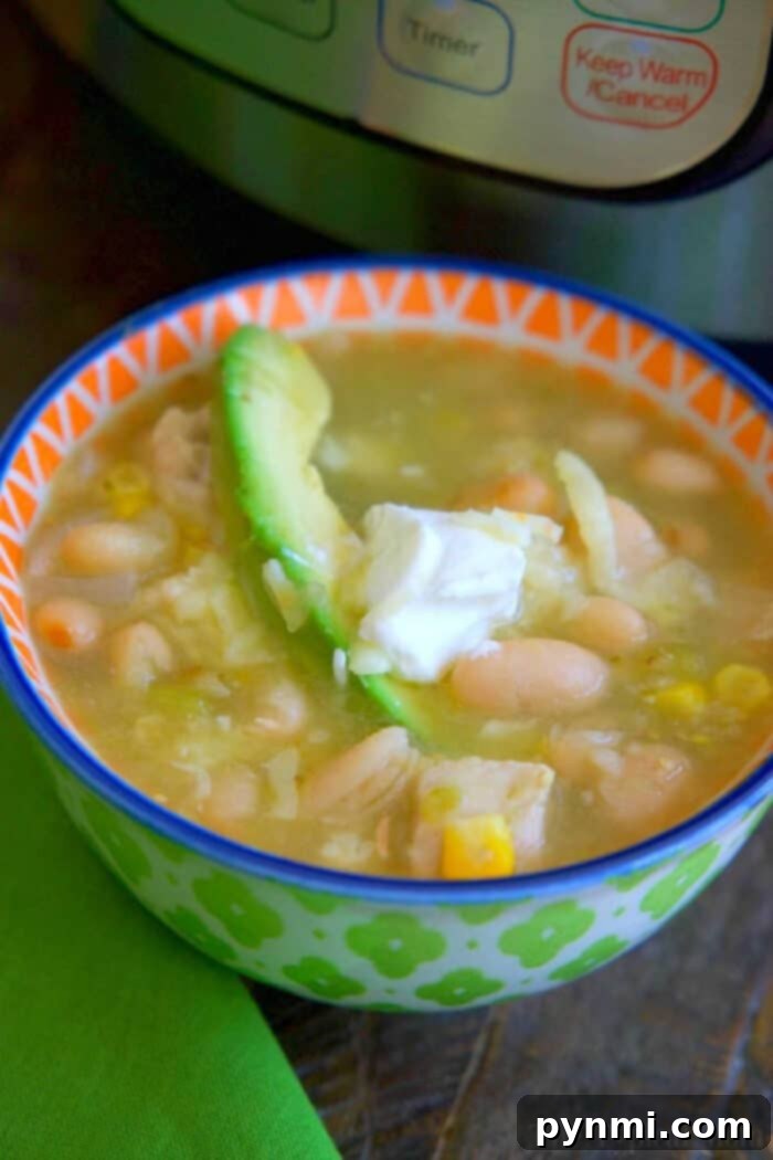A bowl of instant pot white chicken chili topped with avocado and sour cream sits in front of an electric pressure cooker.