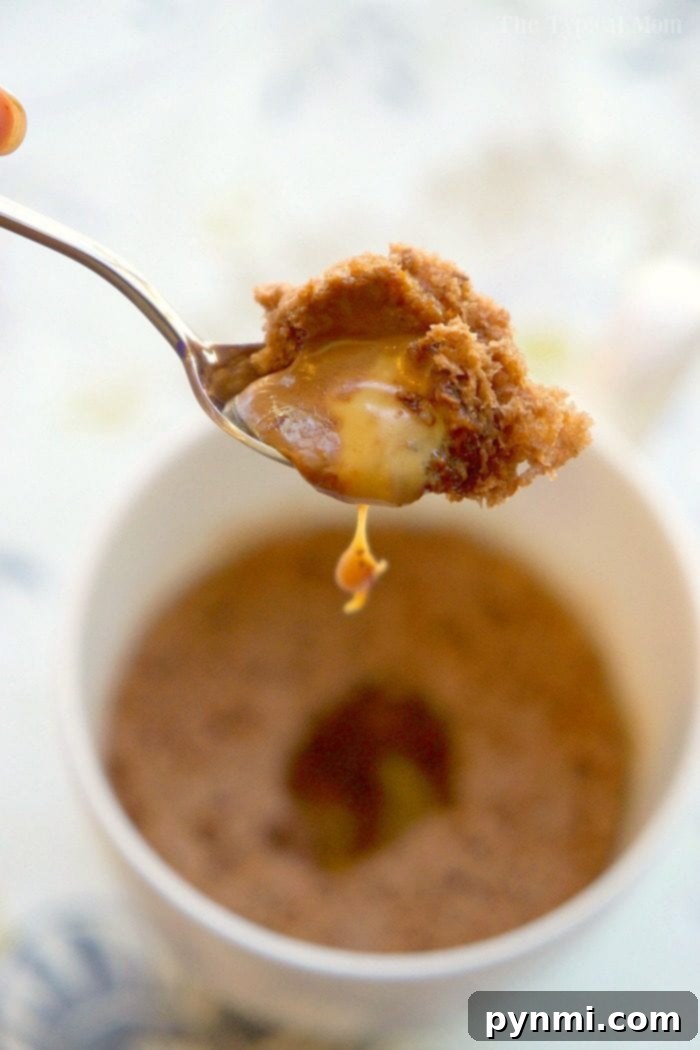 Gooey Caramel Chocolate Mug Cake: A quick and easy microwave dessert with just three ingredients. A spoon delicately lifts a bite of molten caramel and chocolate mug cake, revealing a luscious, gooey center, presented in a crisp white mug on a wooden surface, highlighting the perfect texture.