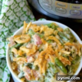 One Pot Wonder Green Bean Casserole 3 A bowl of instant pot green bean casserole topped with crispy onions sits beautifully in front of a pressure cooker, with a green patterned cloth nearby.