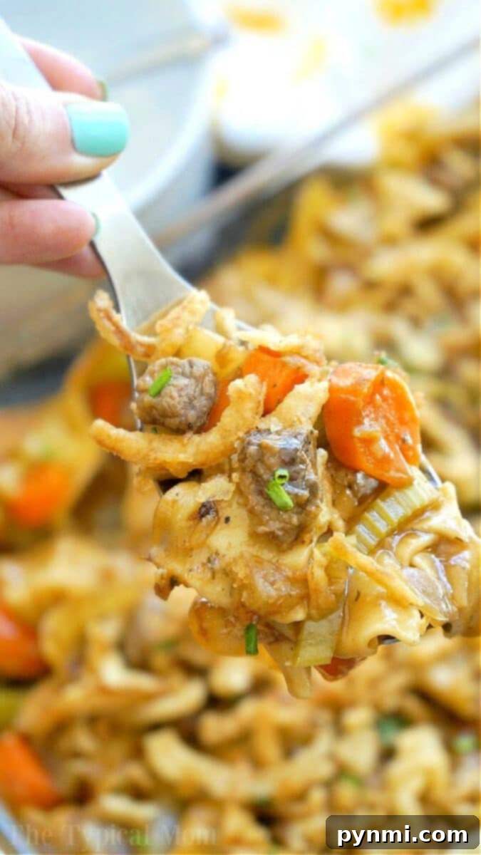 A fork holding a bite of easy beef stew casserole with noodles, beef, carrots, celery, and crispy onions, highlighting the rich texture and vibrant ingredients.