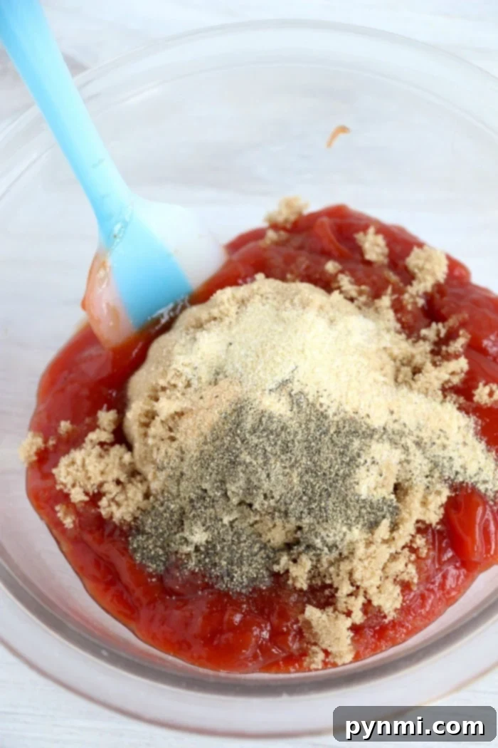 A clear mixing bowl contains ingredients for a savory sauce—ketchup, brown sugar, onion powder, and black pepper. A blue spatula rests in the bowl amid the partially combined mixture.