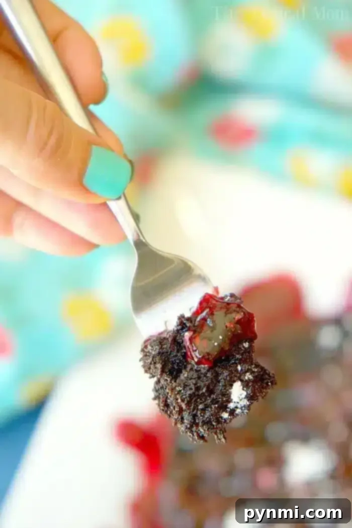 A hand with teal-painted nails holds a fork with a bite of instant pot chocolate cake topped with red berry sauce. In the background, a blurred colorful tablecloth is visible with more of the dessert on a plate, inviting indulgence.