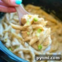 A hand holds a wooden spoon of creamy pasta with green onions over a slow cooker, ready to serve this crockpot pesto chicken alfredo casserole.