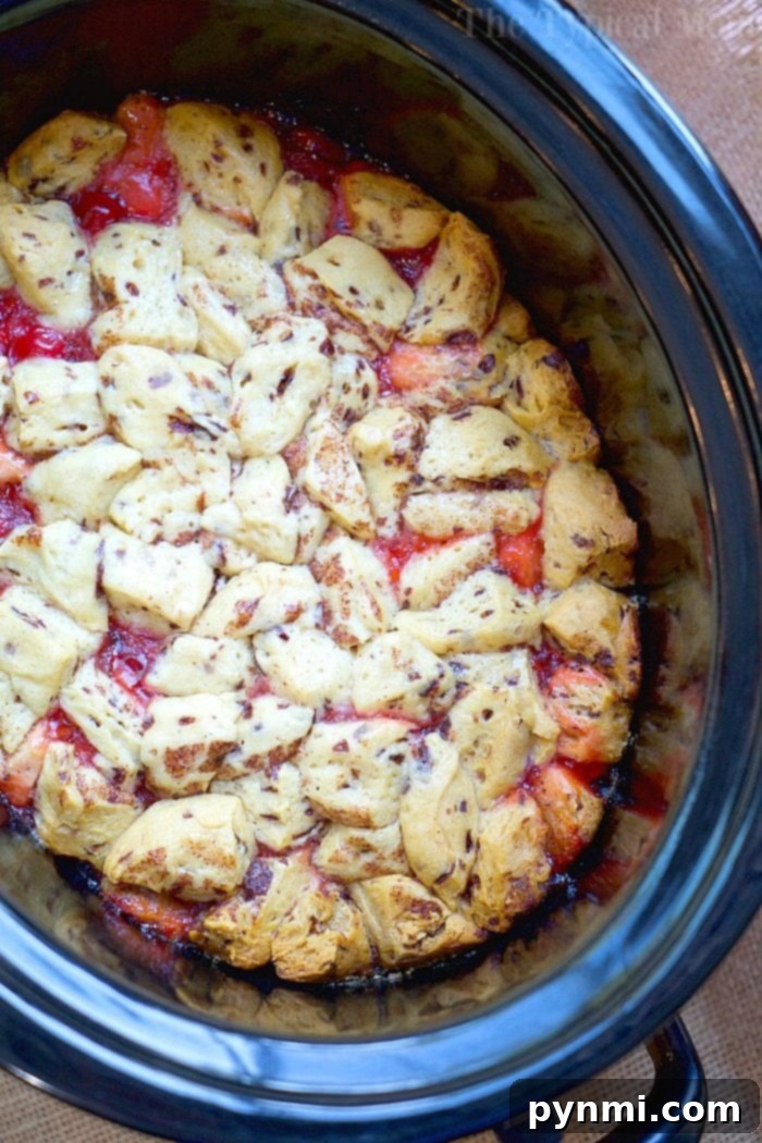 A close-up view of a decadent dessert in a slow cooker. The dish features golden-brown biscuit pieces reminiscent of cinnamon roll monkey bread, resting on a red, fruit-filled base, likely a cobbler. The crockpot has a black rim and sits on a burlap surface.