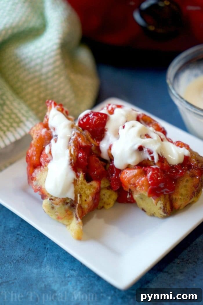 A white plate with two pieces of French toast drizzled in strawberry sauce and cream rests on a blue surface, evoking the coziness of crockpot cinnamon roll monkey bread. In the background, a green and white cloth accompanies a small bowl of additional cream.