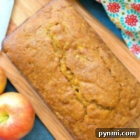 A beautifully golden loaf of easy pumpkin apple bread on a wooden board, garnished with fresh apples and a colorful kitchen towel.