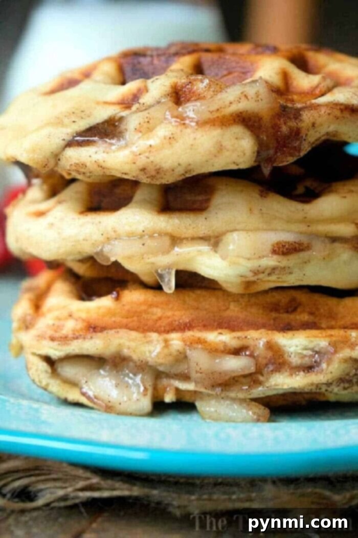 Close-up of a stack of three glazed, apple pie stuffed waffles on a blue plate. The waffles show a golden-brown, slightly crispy texture with a dripping glaze. The background is blurred, putting focus on the cinnamon roll waffles. Text "The Typical Mom" is faintly visible at the bottom right.