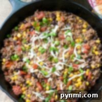 Skillet with ground beef, corn, beans, tomatoes, cheese, and green onions; it’s a taco dip delight paired perfectly with a bowl of tortilla chips in the background.