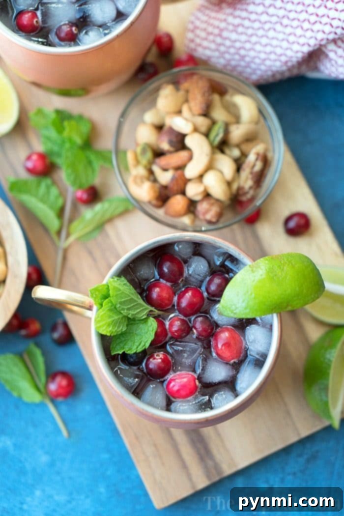 Refreshing Cranberry Moscow Mule: A Festive Holiday Essential. A frosty copper mug filled with a beautiful Cranberry Moscow Mule, adorned with fresh cranberries, a lime wedge, and a sprig of mint, all resting on a rustic wooden board.