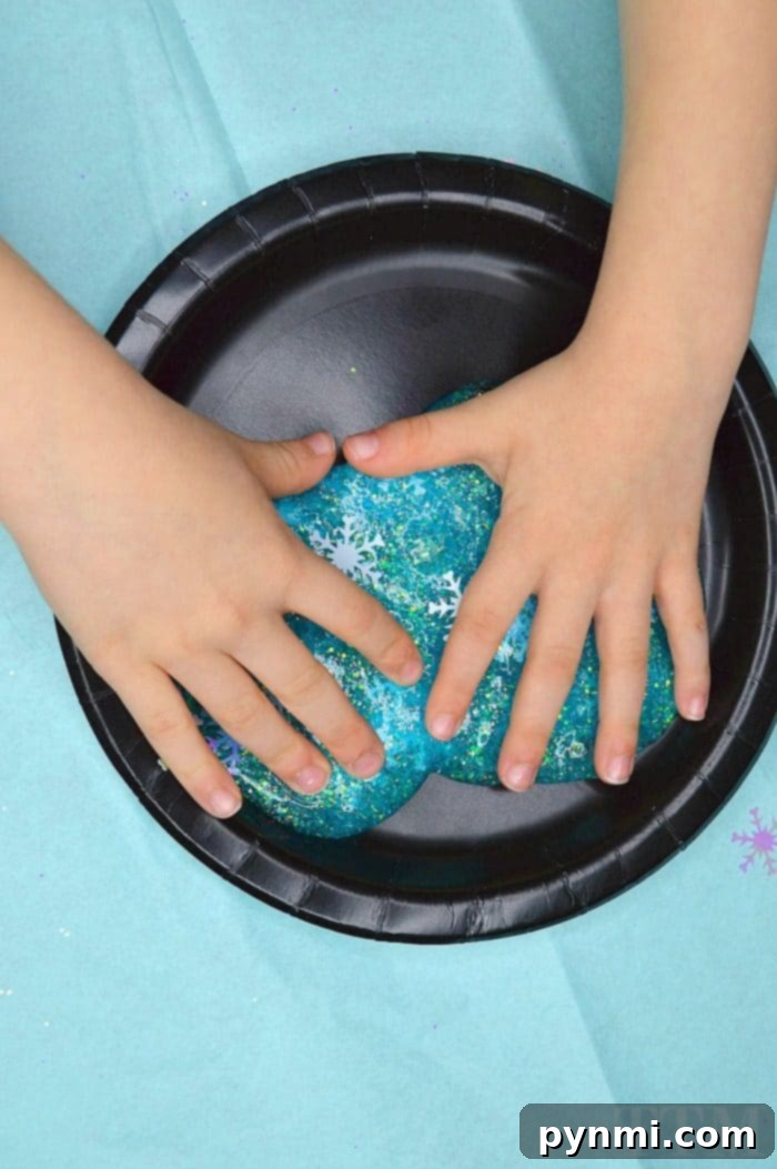 Two hands stretching a beautiful blue winter glitter slime, studded with iridescent snowflakes and fine gold glitter.