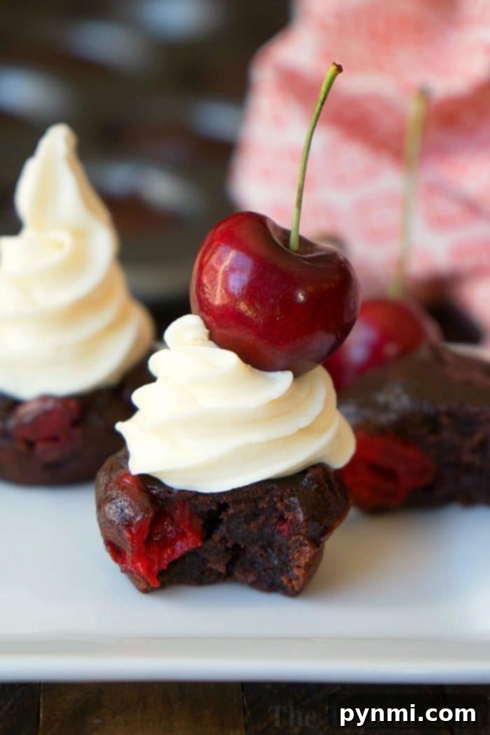 A decadent chocolate cherry brownie, adorned with a swirl of white frosting and a fresh red cherry, sits invitingly on a pristine white plate. In the background, a spread of similar cherry brownies, featuring a rich, dark chocolate interior studded with vibrant red cherries, suggests a tempting array of treats.