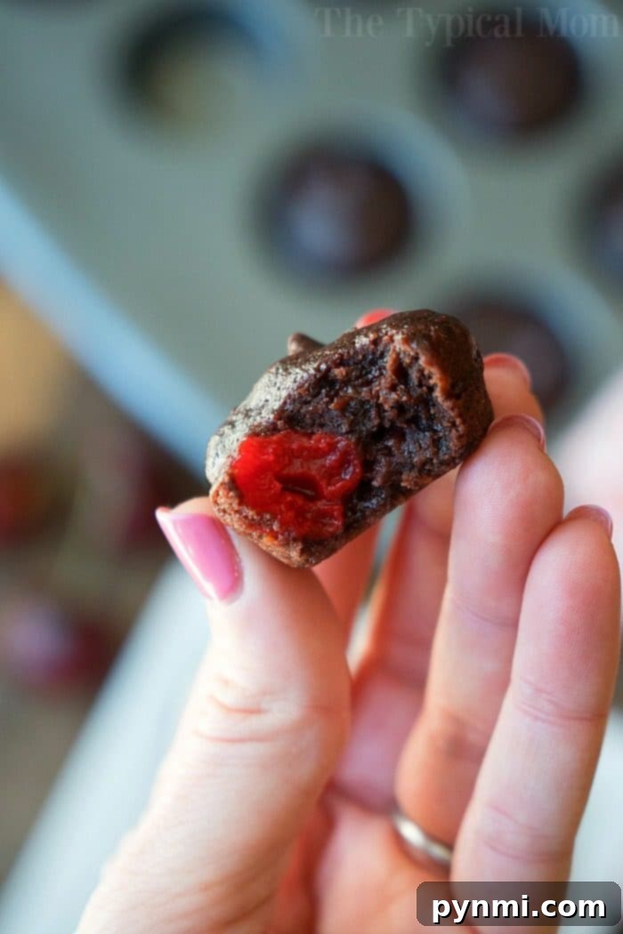 A hand gently holds a small, perfectly baked chocolate brownie, from which a bite has been taken, revealing a bright, luscious red cherry filling. The background is softly blurred, showing a muffin tray filled with more of these inviting cherry brownies, highlighting their individual portions.