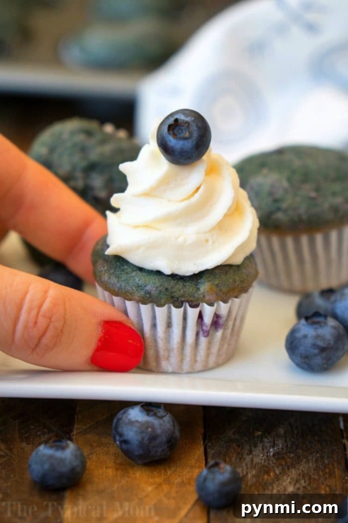 A hand with red nail polish holds a mini blueberry cupcake adorned with white frosting and a fresh blueberry on top. Other cupcakes, scattered blueberries, and hints of blueberry pie filling create a tempting backdrop on a white plate and wooden surface.